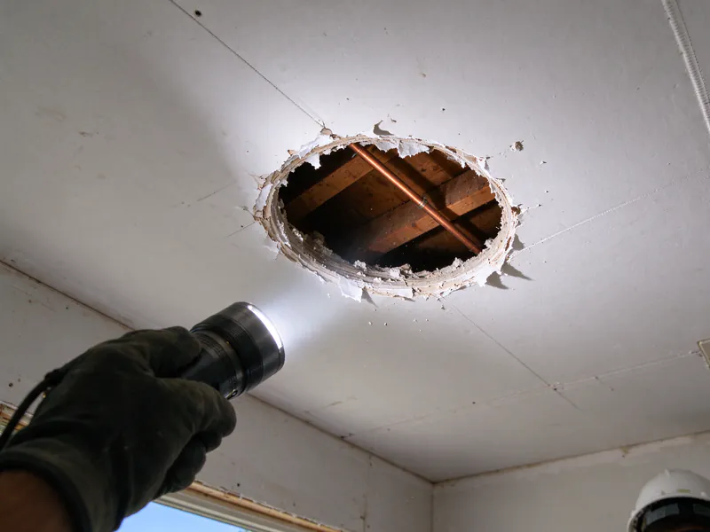 Flashlight shining into a round inspection hole in a ceiling, showing a copper pipe between joists, in an older Columbus home